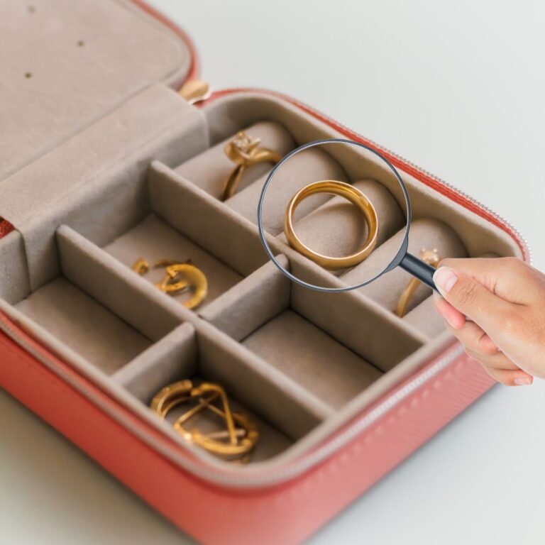 Eine Hand hält eine Lupe, die auf einen goldenen Ring in einem Schmuckkasten zeigt.