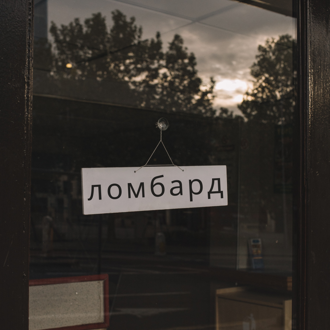 Ein weites Bild einer Scheibe mit einem Schild, das in kyrillischen Buchstaben das Wort 'ломбард' zeigt, hängt an der Tür. Der Hintergrund ist verschwommen mit Baumresten und einem grauen Himmel.